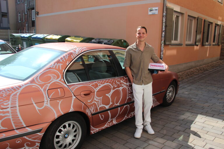 Person steht neben einem farbenfroh gestalteten Fahrschulauto der Fahrschule Cartel und hält ein Schild mit der Aufschrift „Fahrschule“.