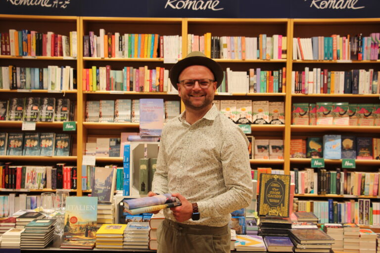 Ein lachender Thalia-Mitarbeiter mit Hut steht vor einer Bücherwand und präsentiert stolz drei Bücher zum Thema Italien.