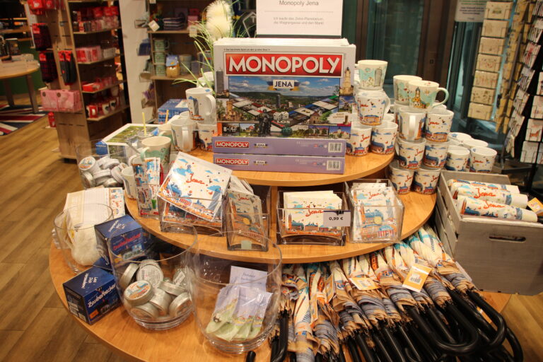 Runder Verkaufstisch mit Jena-Souvenirs wie der Monopoly Jena Edition, bedruckten Tassen, Regenschirmen und Postkarten.