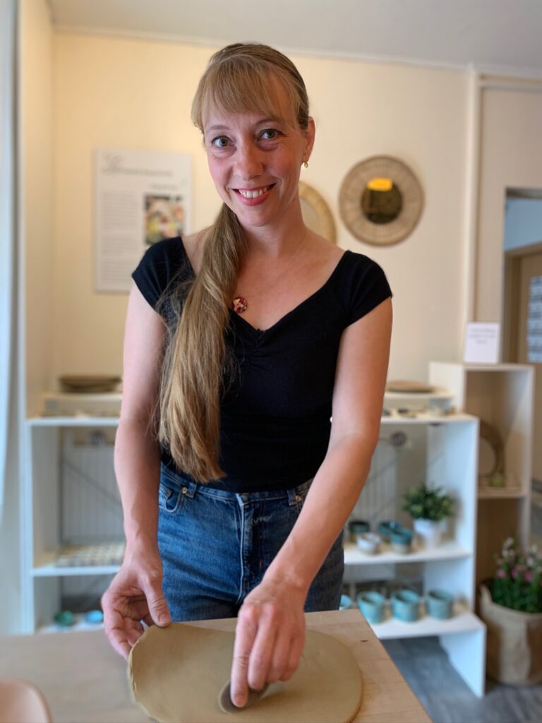 Lächelnde Inhaberin von Jenaceramics steht in ihrer Werkstatt und bearbeitet mit einem Werkzeug eine Tonplatte.