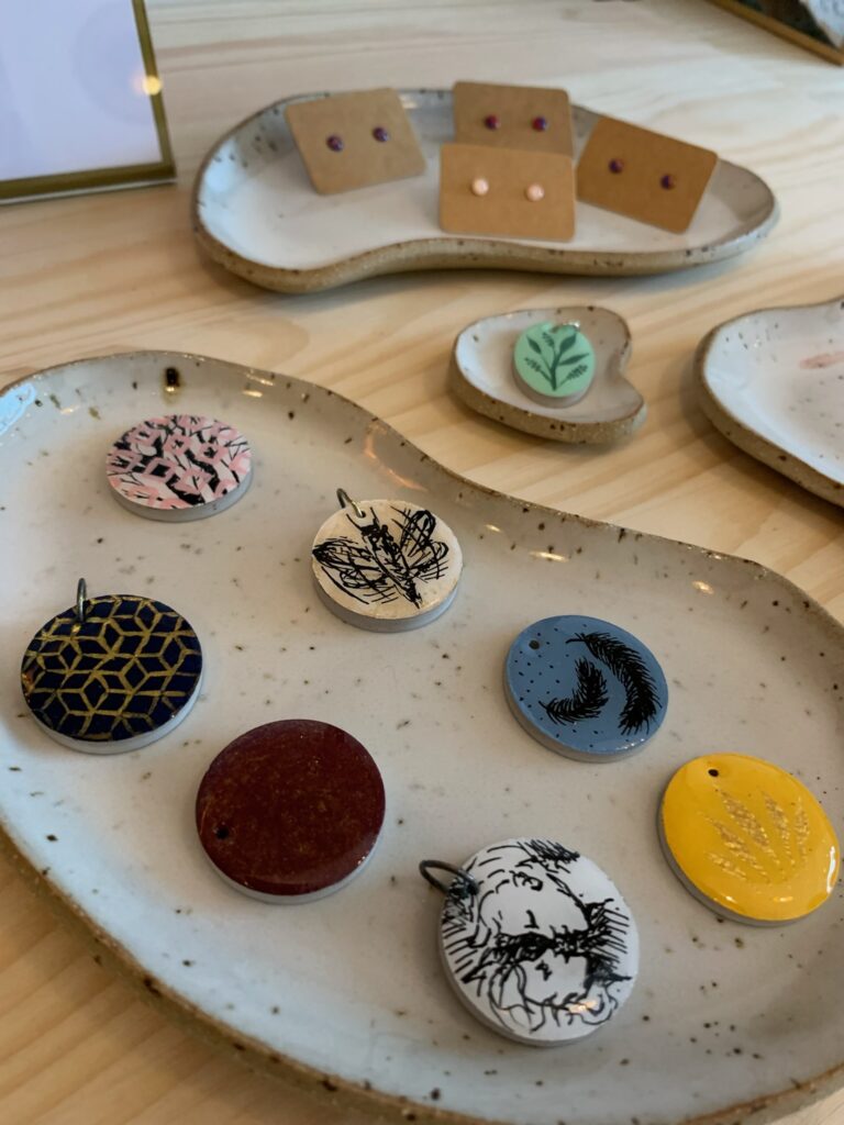 Verschiedene runde Keramik-Anhänger mit handbemalten Motiven wie Blättern, Federn und geometrischen Mustern auf einer Schale.