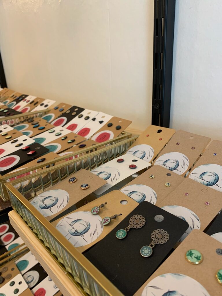 Präsentation zahlreicher kleiner Keramik-Ohrstecker auf Kärtchen in einem gläsernen Kasten, sortiert nach Farben und Mustern.