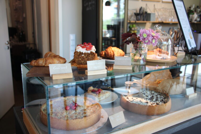 Glasvitrine gefüllt mit hausgemachten Kuchen wie Möhrenkuchen und Käsekuchen, sowie frischen Croissants und Franzbrötchen.