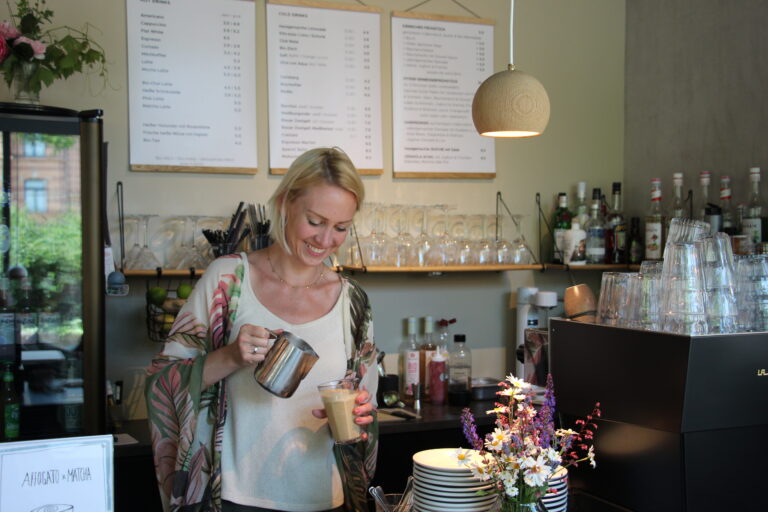Lächelnde Barista gießt Milch in ein Glas hinter der Theke, im Hintergrund eine helle Espressomaschine und Menütafeln.