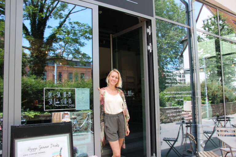 Die Inhaberin steht freundlich lächelnd in der geöffneten Glastür des Holz & Hygge Stores, auf der die Öffnungszeiten stehen.