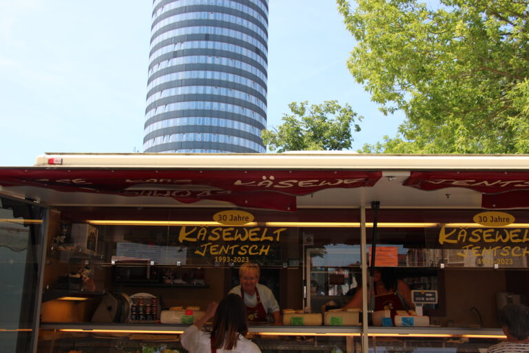 Der rot-weiße Verkaufswagen der „Käsewelt Jentsch“ auf dem Marktplatz, im Hintergrund ragt der moderne Jentower in den blauen Himmel.