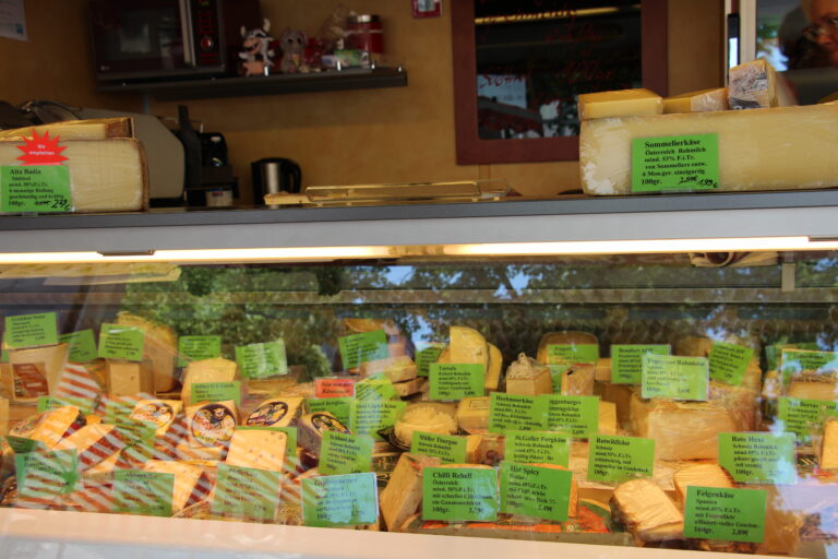 Nahaufnahme der gut gefüllten Käsetheke mit verschiedenen Sorten wie Sommelierkäse, Alta Badia und Chili-Käse, ausgezeichnet mit grünen Preisschildern.