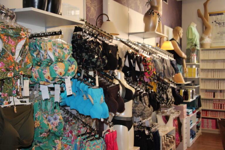 Gut sortierte Kleiderständer mit bunter Bademode, Bikinis und Badeanzügen mit tropischen Mustern; im Hintergrund Mannequins.