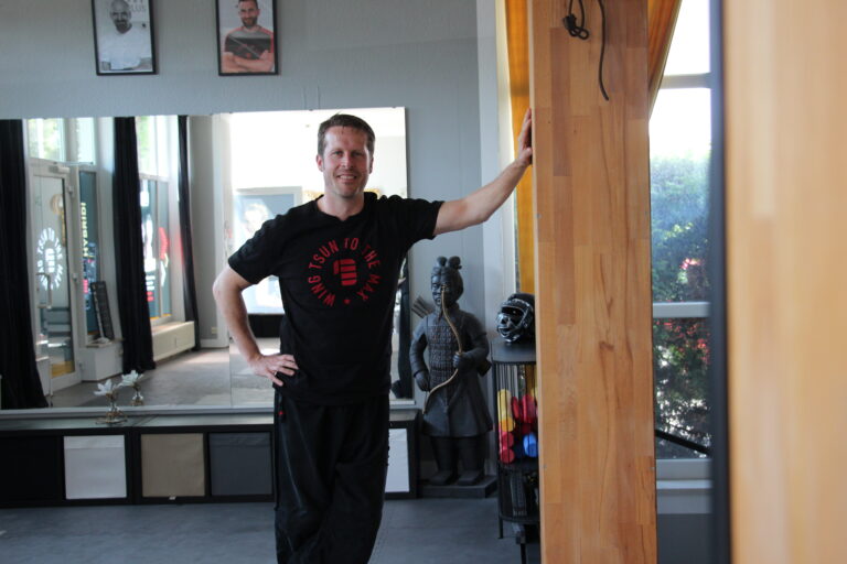 Der Schulleiter steht lächelnd und entspannt in schwarzer WingTsun-Kleidung an einer Holzsäule im Trainingsraum.