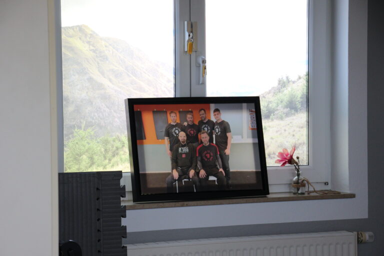 Gerahmtes Gruppenfoto von sechs WingTsun-Trainern in Uniform auf einer Fensterbank, mit Blick ins Grüne im Hintergrund.
