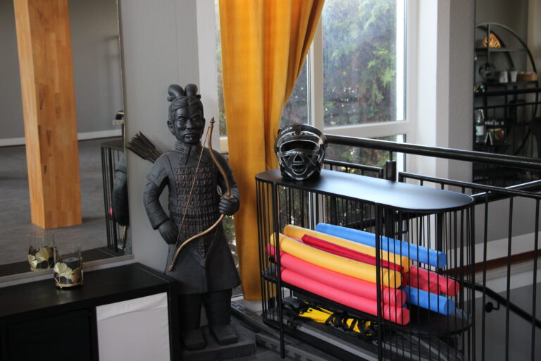 Eine schwarze Terrakotta-Krieger-Statue mit Bogen steht neben einem modernen Regal mit Kopfschutz und bunten Soft-Sticks.