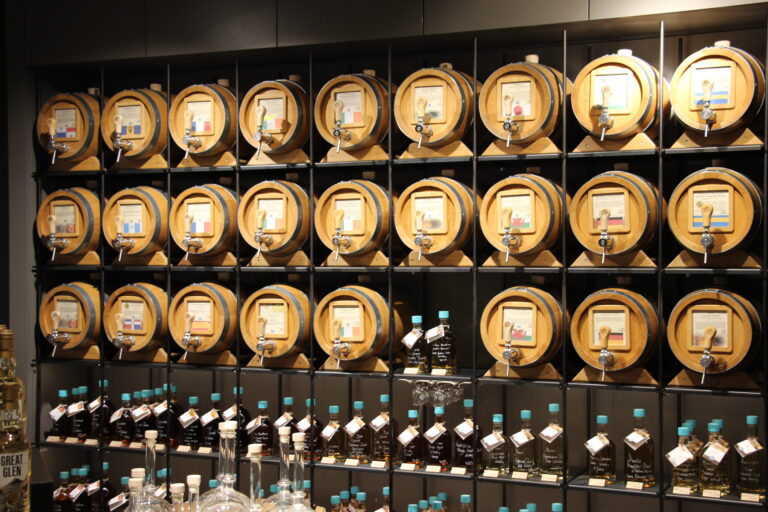 Wandregal mit vielen kleinen Holzfässern mit Zapfhähnen, beschriftet mit verschiedenen Whisky- und Rumsorten und Länderflaggen.