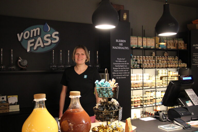 Eine freundliche Mitarbeiterin steht hinter dem Tresen unter schwarzen Pendelleuchten, neben ihr ein Schild mit der Aufschrift "Bleiben Sie nachhaltig".