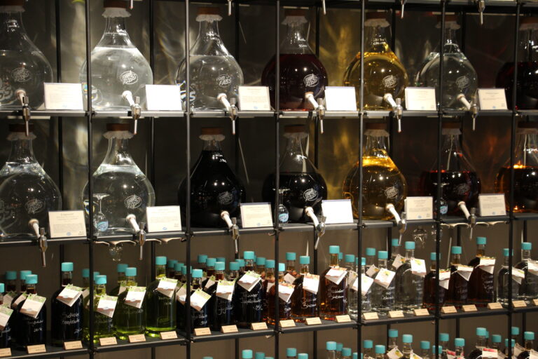 Alternativtext: Regalwand mit großen, runden Glasballons (Demijohns), gefüllt mit klaren und dunklen Flüssigkeiten wie Essigen und Ölen, darunter abgefüllte Flaschen.