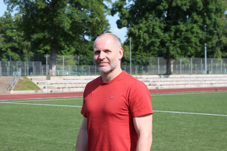 Porträt eines Mannes im roten T-Shirt, der freundlich lächelnd auf einem Sportplatz vor grünen Bäumen steht.