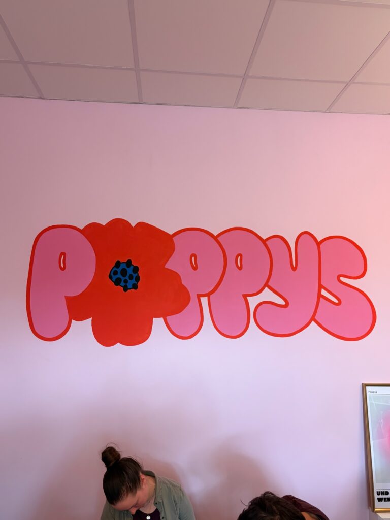 Wand mit dem handgemalten Poppys-Logo in Rot und Rosa, darunter Personen bei einem Workshop.
