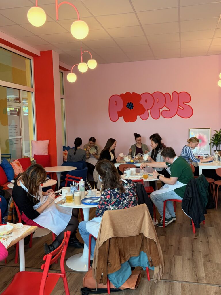 Mehrere Menschen sitzen in einem farbenfrohen Café und bemalen Keramik an kleinen Tischen.