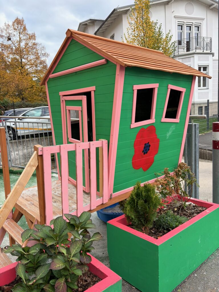 Kleines, grün-rosafarbenes Holz-Spielhaus mit schrägen Fenstern und rotem Blütenmotiv, umgeben von bepflanzten Hochbeeten.