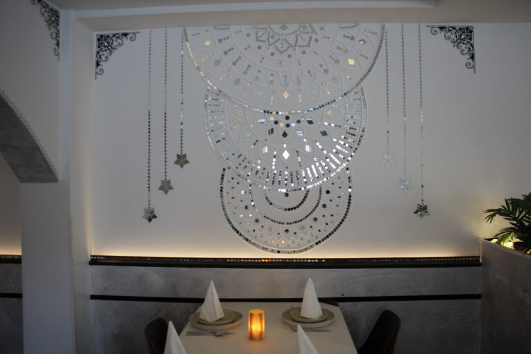 Dekorative Wand im Naan n Curry mit glänzenden Spiegelmosaiken in Mandala-Form über einem gedeckten Tisch.