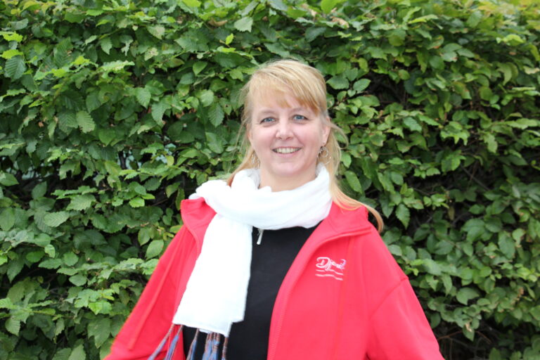 Porträt von Djamila, der Erfolgsmentorin, freundlich lächelnd in roter Jacke und weißem Schal vor einer grünen Hecke im Freien.