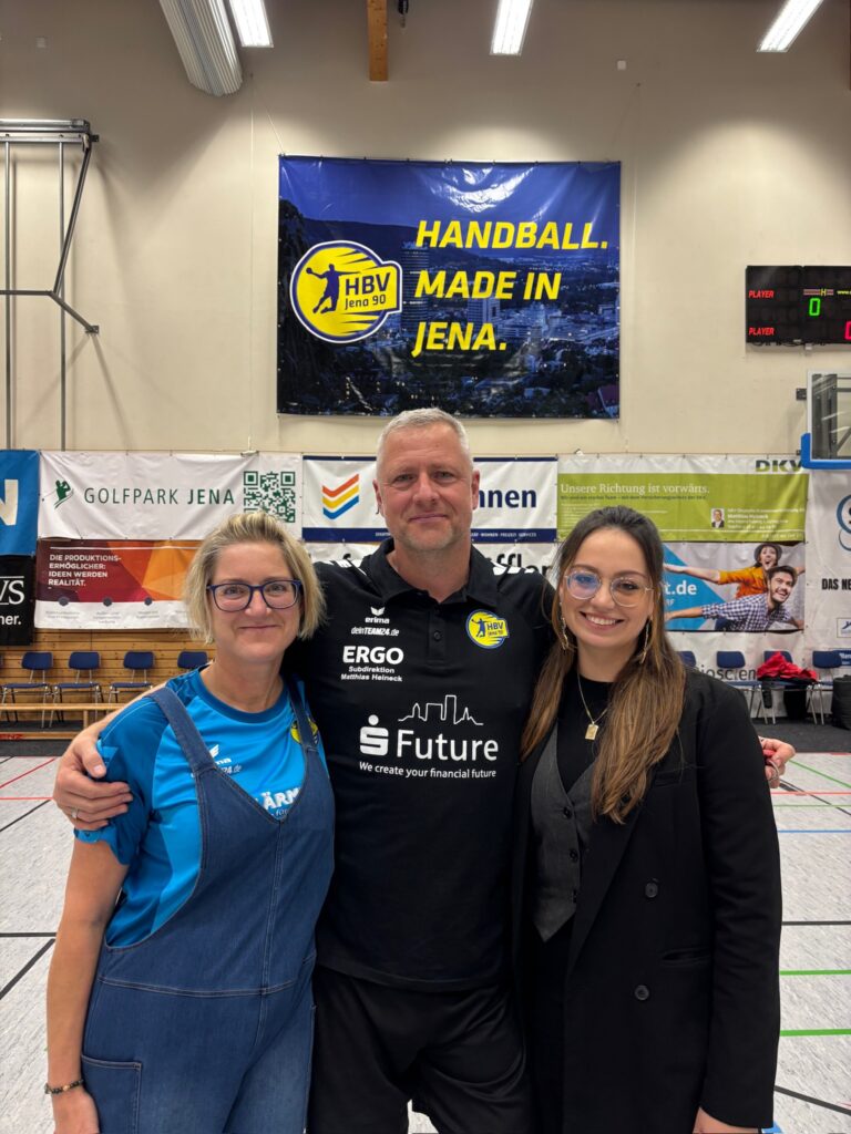 Drei Vereinsmitglieder des HBV Jena stehen lächelnd Arm in Arm vor einem großen Banner mit der Aufschrift "Handball. Made in Jena.".