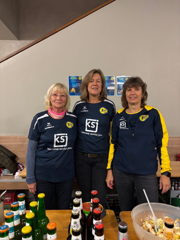 Drei freundliche Damen vom Verpflegungsteam stehen in Vereinskleidung hinter einem Tresen mit Getränken und Obstsalat.