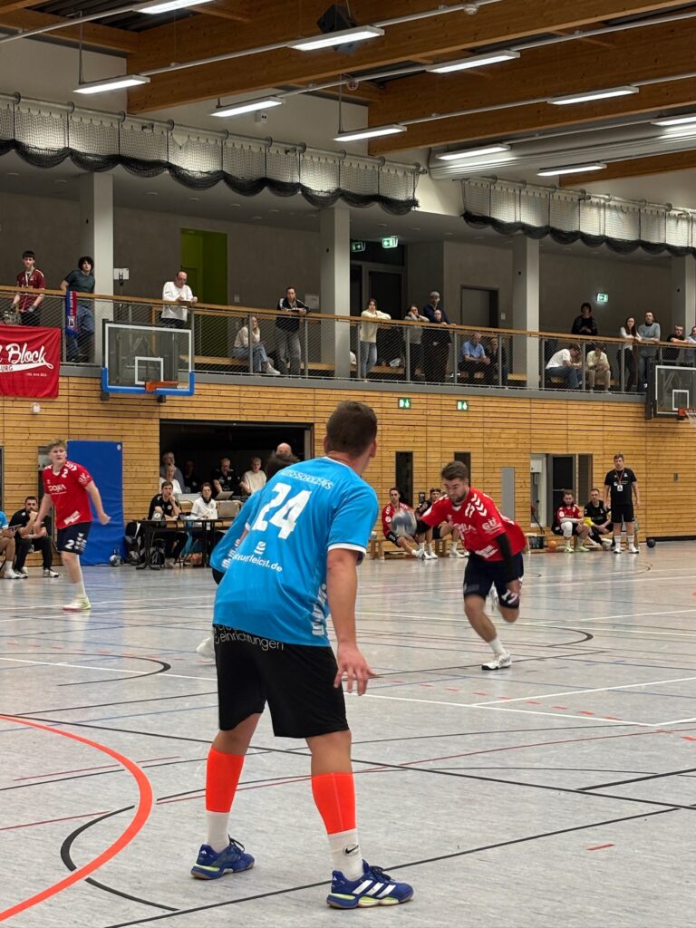 Dynamische Spielszene beim Handballspiel: Ein Spieler des HBV Jena in Blau verteidigt gegen einen Angreifer in Rot vor gefüllten Zuschauerrängen.