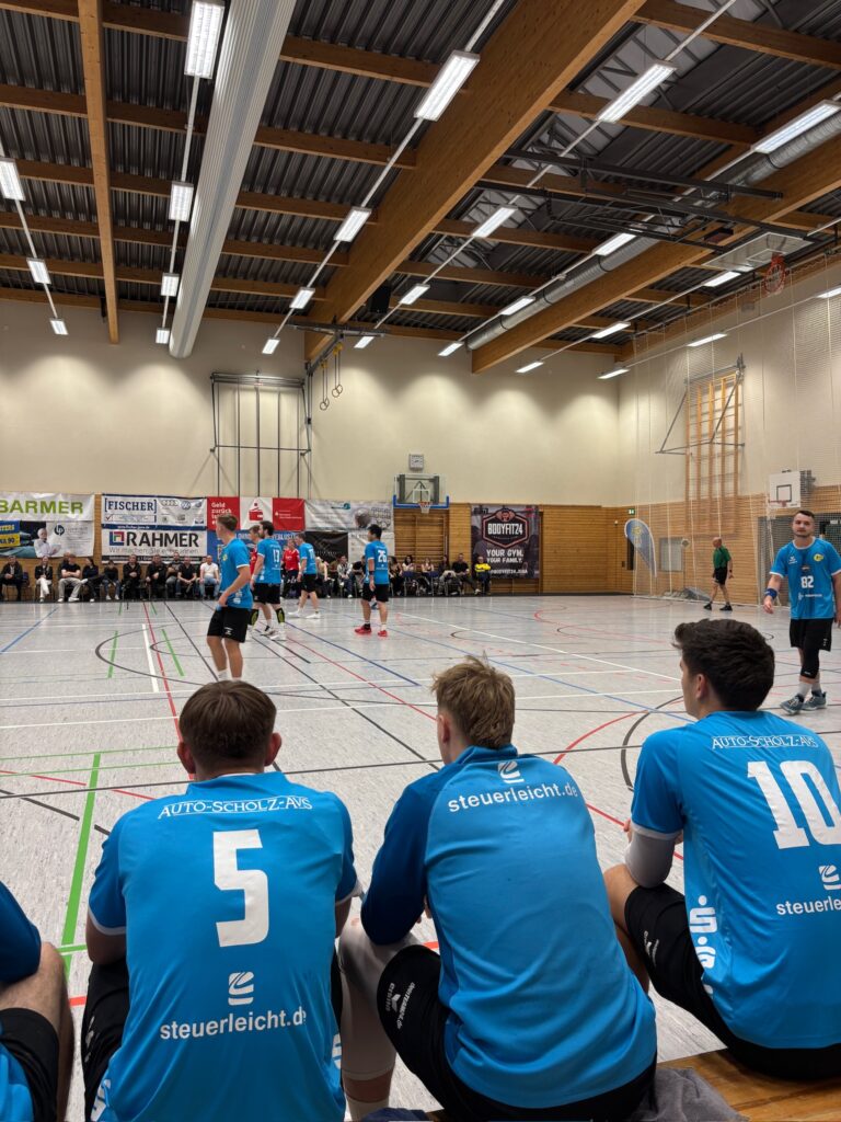 Handballspieler des HBV Jena in blauen Trikots sitzen auf der Auswechselbank und beobachten gespannt das Spiel in der Sporthalle.