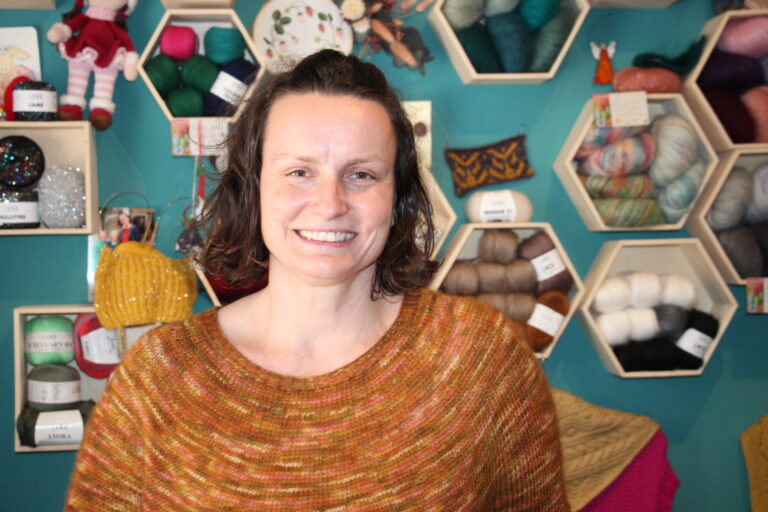 Porträt der freundlichen Inhaberin von Fadenwohl vor einer Wanddekoration mit Wollregalen in Wabenform.