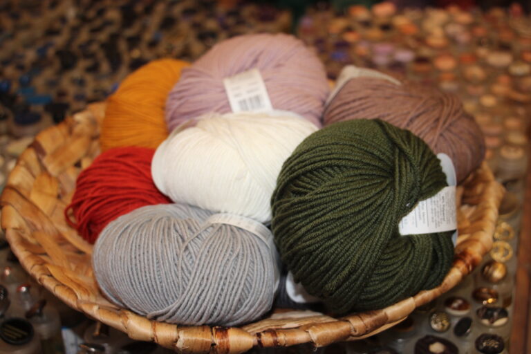 Eine Auswahl von Wollknäueln in verschiedenen Unifarben (Grau, Grün, Weiß, Lila) in einem geflochtenen Korb.