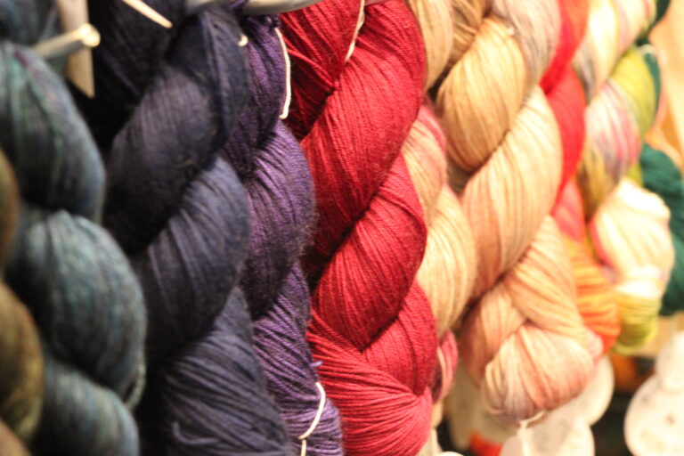 Nahaufnahme von hängenden, in Strängen gewickelten Garnen in leuchtenden Farben, darunter dunkles Blau, Rot und Beige-Töne.