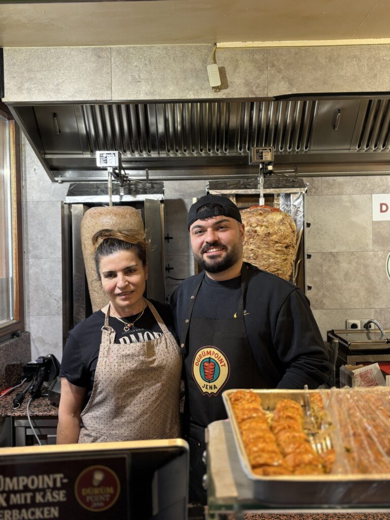 Inhaber:innen von Dürüm Point Jena stehen lächelnd in Schürzen vor den Döner-Spießen hinter der Theke.