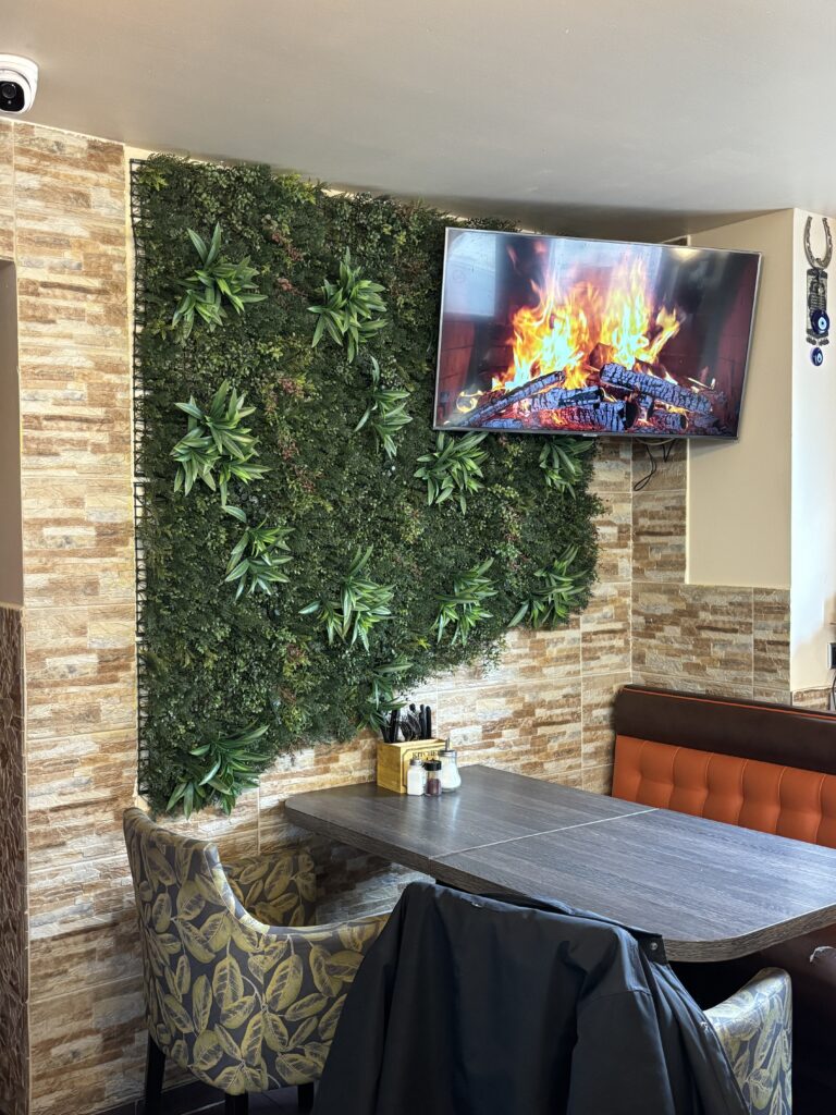 Gemütliche Sitzecke im Dürüm Point mit gepolsterten Bänken, Pflanzenwand und einem digitalen Kaminfeuer auf dem Bildschirm.