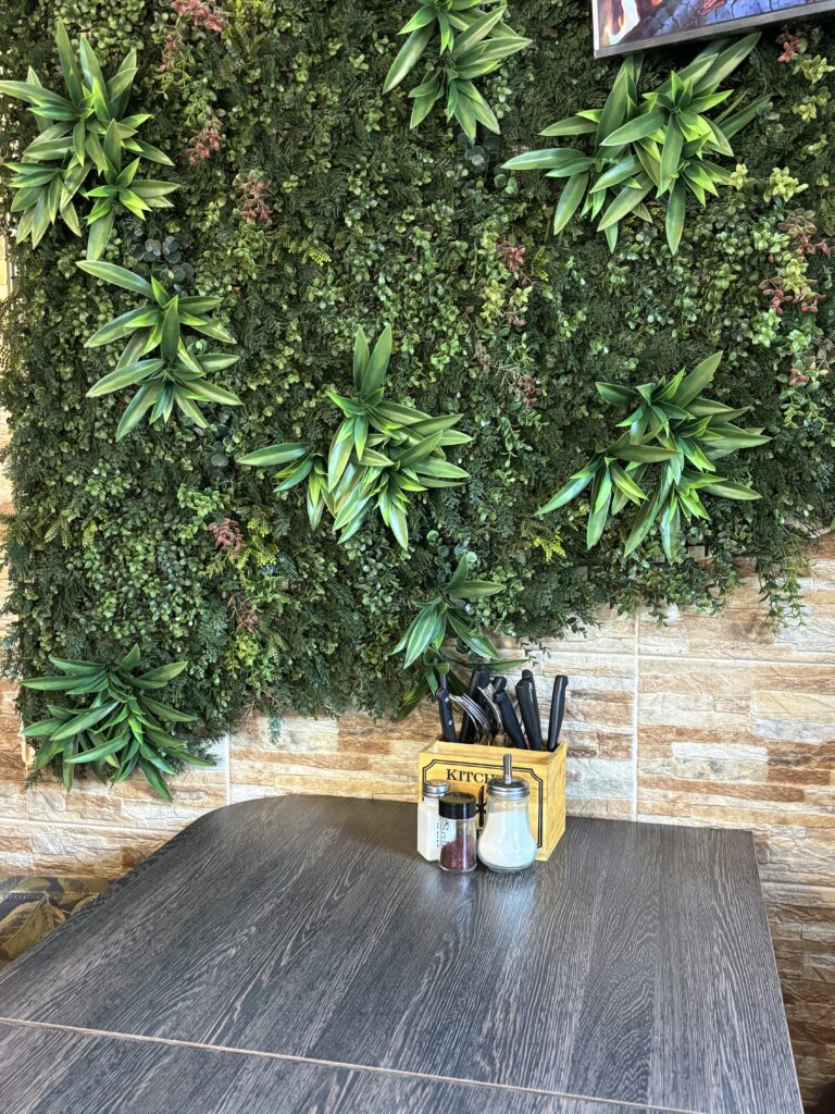 Tisch mit Besteck und Gewürzen vor einer dekorativen Wand aus künstlichen Pflanzen im Dürüm Point Jena.