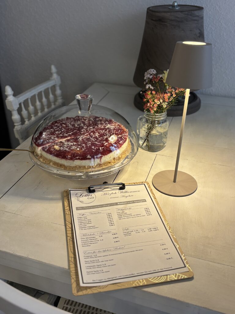 Hausgemachter Käsekuchen unter einer Glashaube und Menükarte auf einem weißen Holztisch im Lotti's Café Jena.