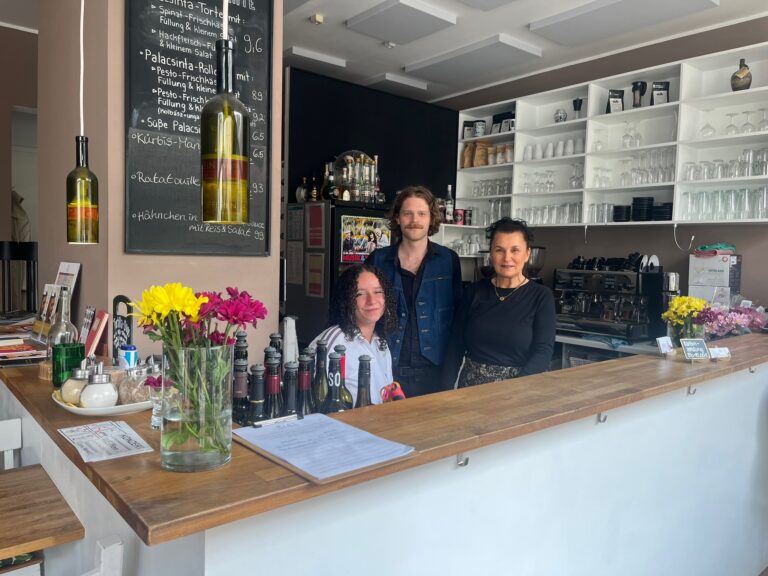 Das freundliche Team vom Pici hinter dem Holztresen, im Hintergrund eine Tafel mit ungarischen Spezialitäten und Wein-Lampen.