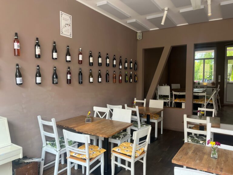 Gemütlicher Gastraum im Pici mit weißen Stühlen, bequemen Sitzkissen und einer dekorativen Wandinstallation aus Weinflaschen.