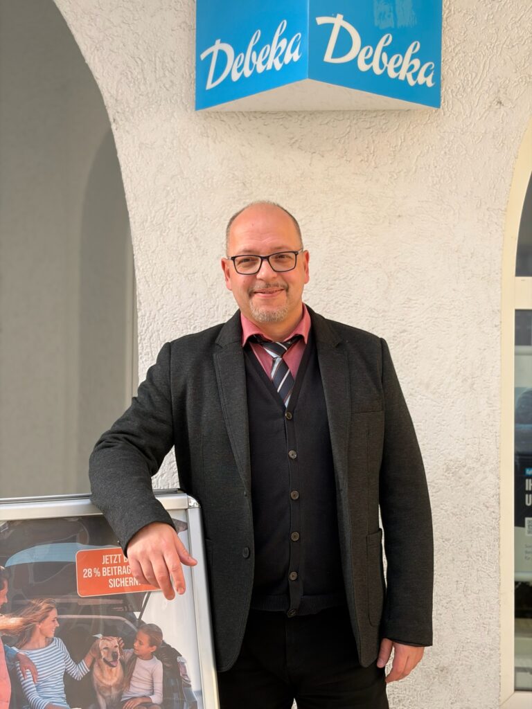 Teamleiter von Debeka mit Brille und Sakko steht neben einem Werbeaufsteller vor der Bürotür in Jena.