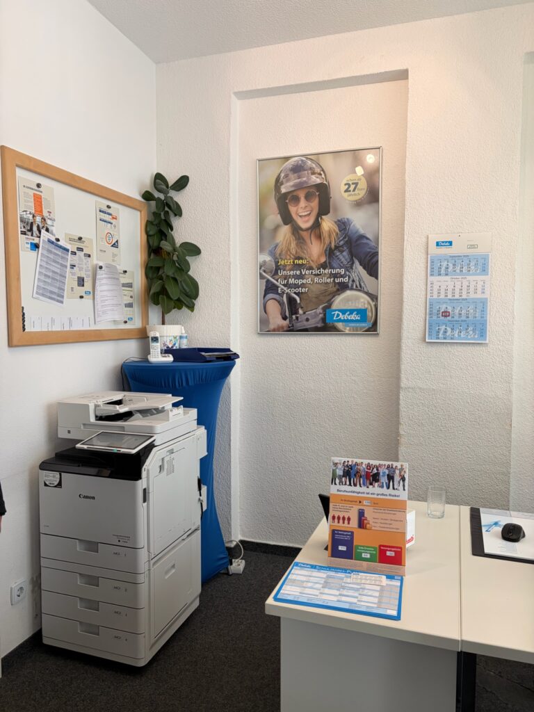 Büroecke mit großem Drucker, Zimmerpflanze und einem Poster für Moped-, Roller- und E-Scooter-Versicherung an der Wand.