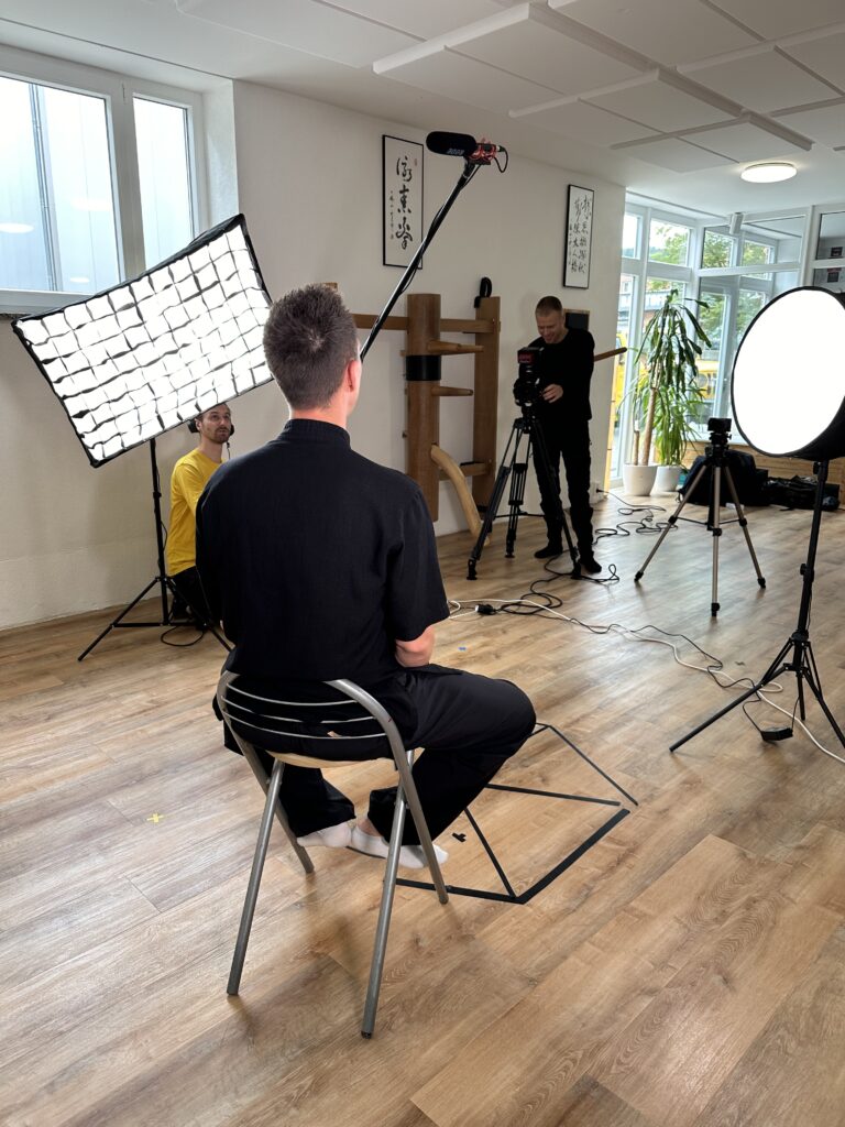 Making-of-Szene eines Interview-Drehs in einem hellen Raum: Ein Kameramann filmt einen sitzenden Interviewpartner, ausgeleuchtet von großen Softboxen, während der Ton über eine Angel geangelt wird.