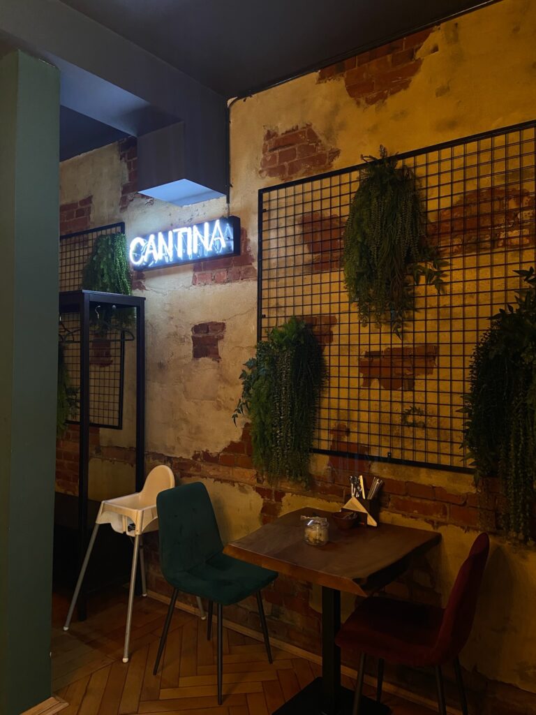 Ein blauer Neon-Schriftzug mit der Aufschrift „CANTINA“ an einer rustikalen Backsteinwand, ergänzt durch schwarze Gitterelemente und Grünpflanzen.