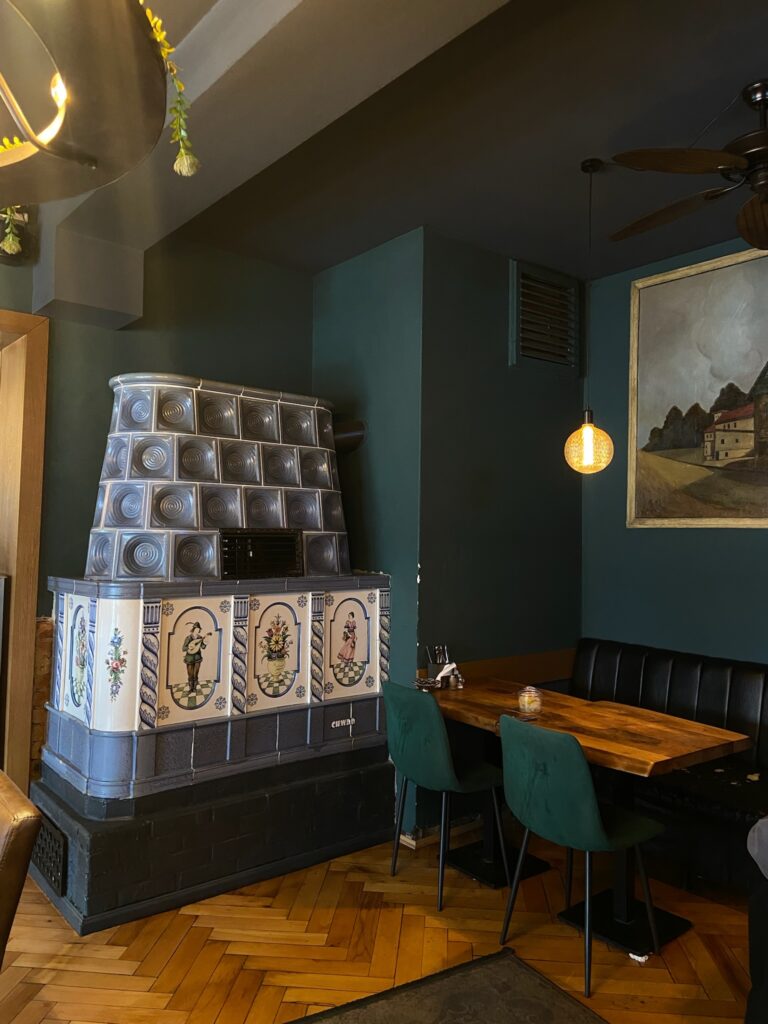 Ein kunstvoll verzierter, blauer Kachelofen neben einem Holztisch mit grünen Samtsesseln in einem dunkelgrün gestrichenen Gastraum.