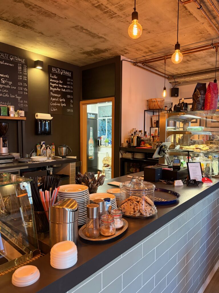 Blick auf den modernen Verkaufstresen im Industrial-Style mit grauen Fliesen, einer gefüllten Gebäckvitrine, Gläsern mit hausgemachten Cookies und warmen Hängelampen.
