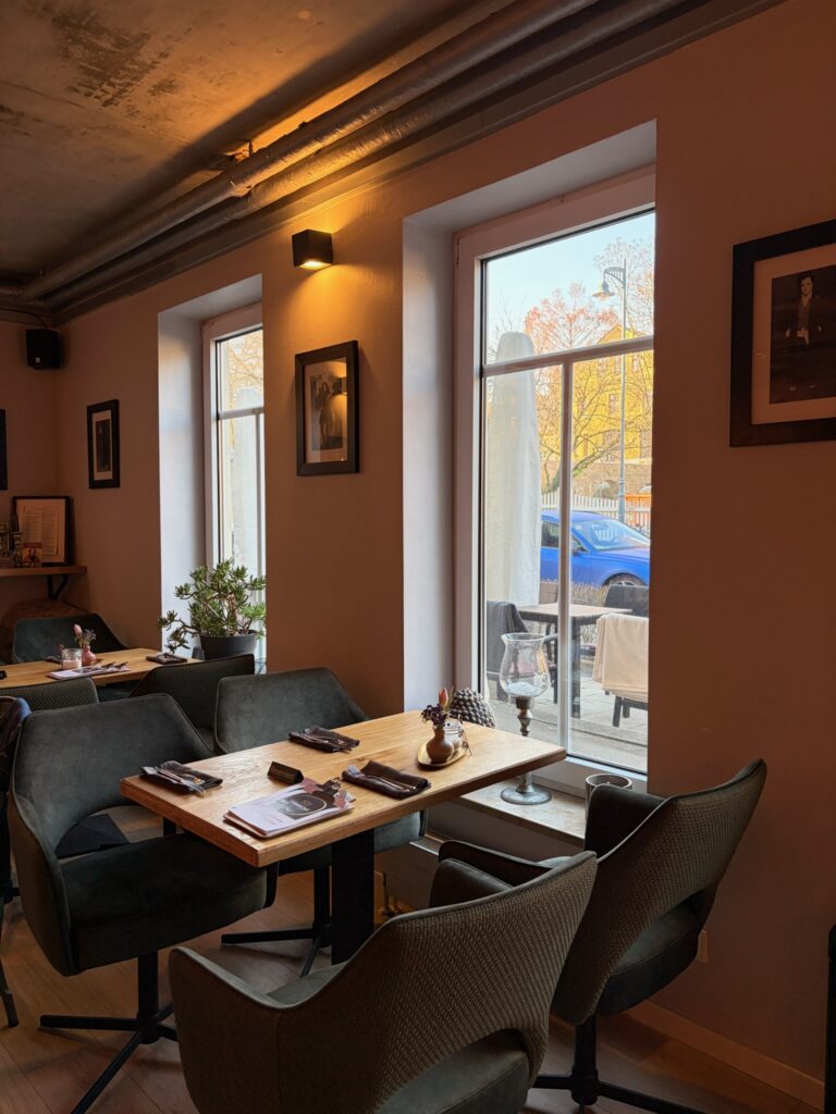 Gemütlicher Gastraum mit bequemen grauen Polstersesseln an Holztischen, großen Fenstern zur Straße und stilvoller Dekoration auf den Tischen