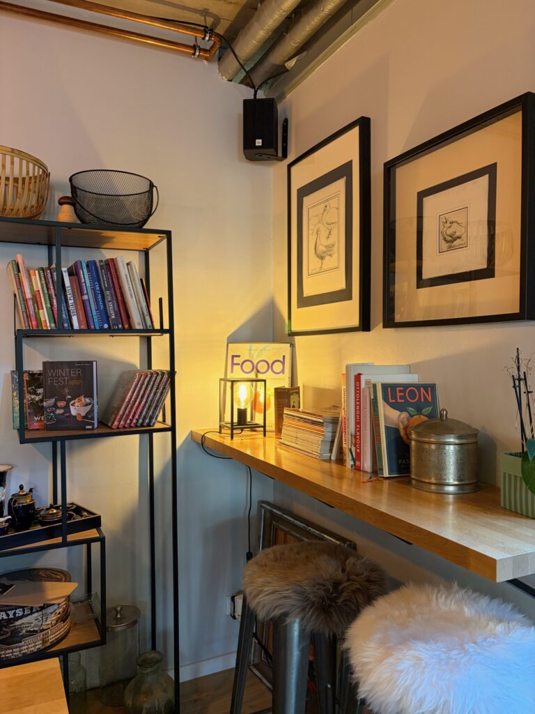 Eine gemütliche Ecke mit warmem Licht, Fellhockern und einem Regal voller Kochbücher (z.B. Ottolenghi, Leon) sowie Romane.