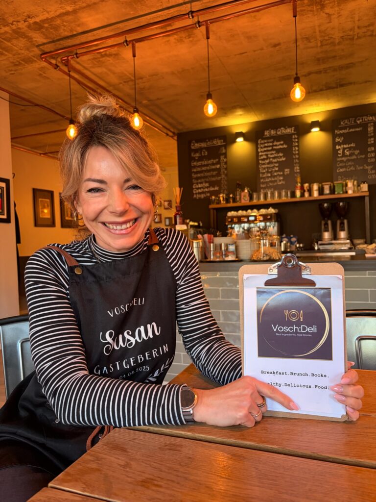Inhaberin Susan sitzt strahlend lächelnd an einem Holztisch, trägt eine Schürze mit der Aufschrift „Susan Gastgeberin 01.08.2025“ und zeigt auf die Speisekarte mit dem Vosch:Deli-Logo.