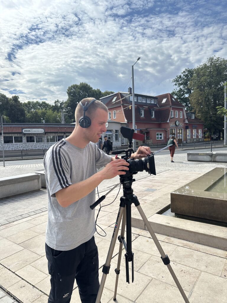 Ein Videograf steht konzentriert an seiner Kamera auf einem öffentlichen Platz in Jena, trägt Kopfhörer und justiert die Einstellungen am Objektiv.