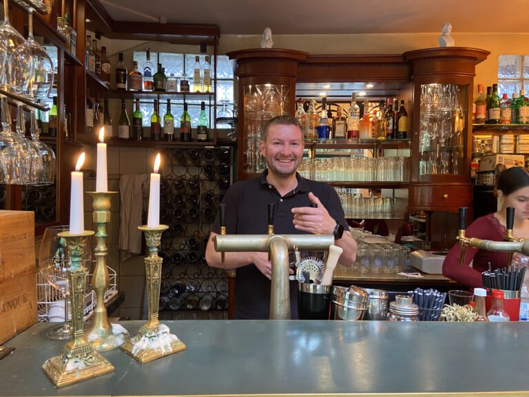 Ein freundlich lachender Barkeeper steht hinter dem Tresen mit Zapfhähnen und Kerzenständern; im Hintergrund gut gefüllte Regale mit Spirituosen.