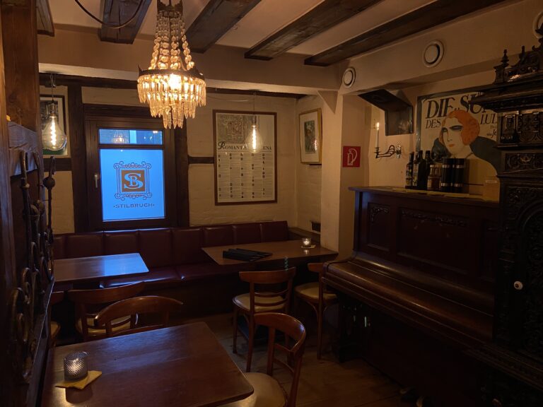 Gemütliche Ecke mit einem Klavier, einem antiken Kristallleuchter an der Decke und einem Jugendstil-Poster an der Wand; im Fenster leuchtet das blaue "SB"-Logo.