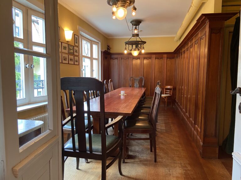 Blick in einen separaten Gastraum mit edler Holzvertäfelung an den Wänden, einem langen Holztisch mit vielen Stühlen und antiken Hängelampen.